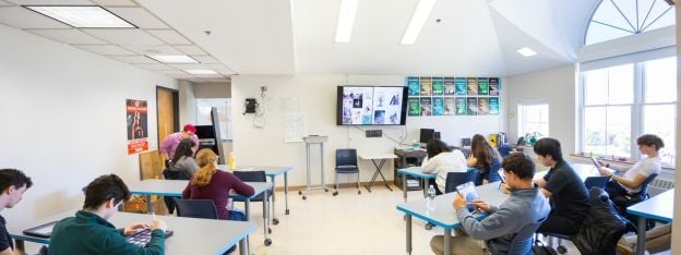 Students sit at desks in a bright classroom, facing a screen at the front. Some write or read, while an instructor stands near a whiteboard. Large windows let in natural light.