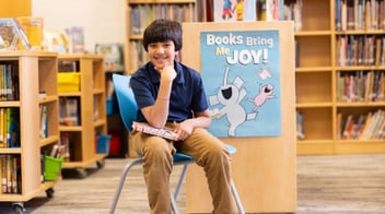 A young boy sits on a chair in a library, holding a book and smiling. Behind him is a sign with cartoon animals that says, "Books Bring Me Joy!" Bookshelves fill the background.