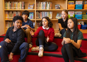 Five children sit on steps in a library, with bookshelves behind them. One child in a red sweater gestures while talking, and the others listen or look thoughtful.