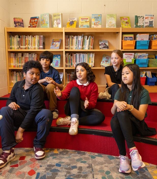 Five children sit on red carpeted steps in a library, with bookshelves behind them. One girl in a red sweater is speaking while the others listen or look on.