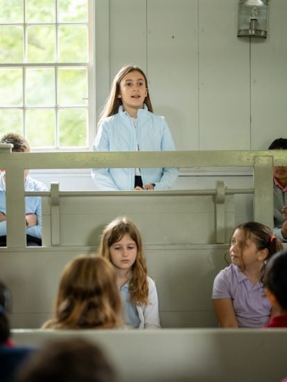 A girl is standing and speaking inside a classroom with several other children seated on wooden benches around her.