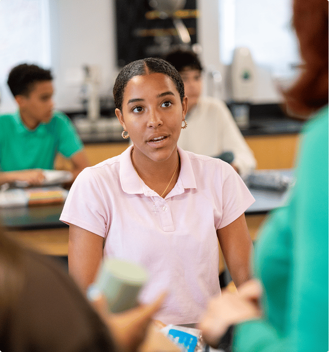 A student in a pink polo shirt listens attentively to a teacher in a classroom, with other students visible in the background.