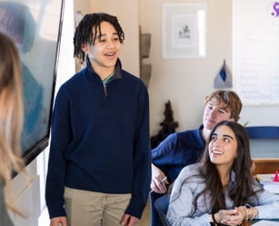 A student stands and speaks at the front of a classroom while classmates sit and listen attentively.