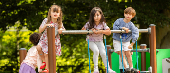 Four young children play on playground equipment outdoors, with two girls and one boy standing on a platform and one child leaning over, facing away. Trees are visible in the background.