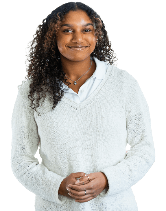 A person with curly hair, wearing a white collared shirt under a light sweater, stands facing the camera and smiles with hands clasped.