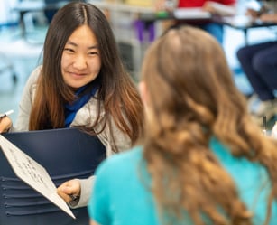 Two students sit facing each other in a classroom. One is holding a whiteboard and smiling, while the other is turned away from the camera.