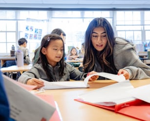 An adult and a child sit at a classroom table, looking at papers together. Other children and classroom materials are visible in the background.