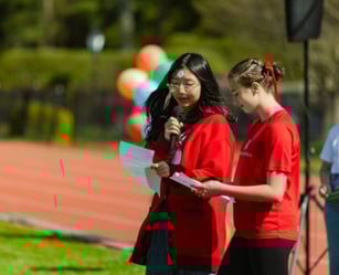 Two young people stand outdoors on a track, holding papers and a microphone, with colorful balloons and blurred figures in the background.
