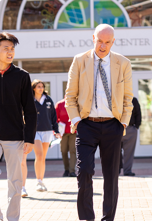 A man in a beige blazer and tie walks outside with hands in his pockets, accompanied by a younger man and several people in the background near a building entrance.