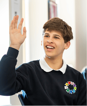 A teenage boy wearing a black sweater with a colorful emblem raises his hand while seated indoors.