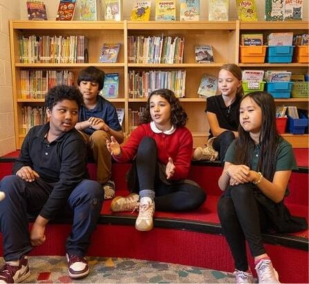 Five children sit on red steps in a library, with bookshelves and colorful bins in the background. One girl in a red sweater is speaking while others listen.