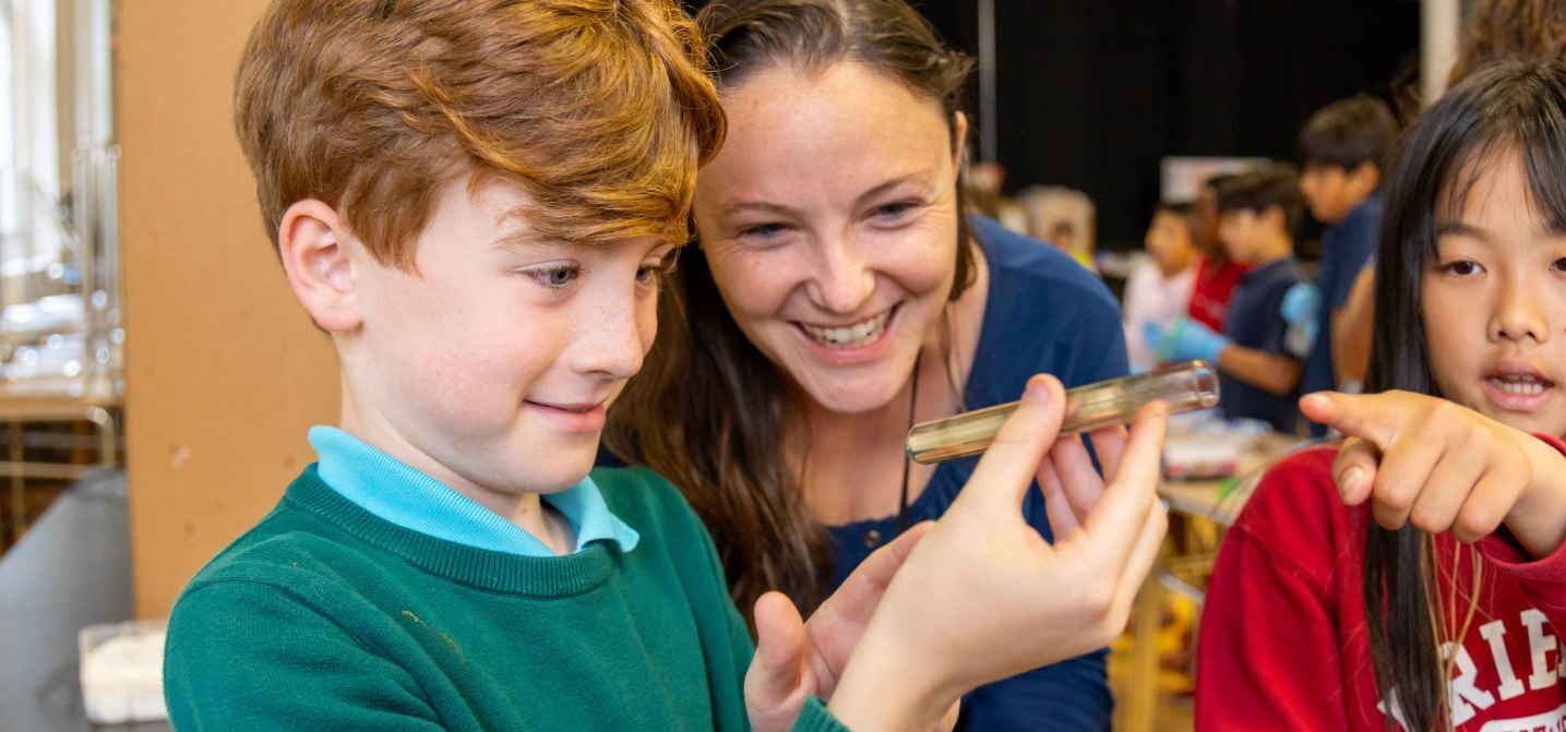 A boy examines a glass slide while a woman smiles behind him and another child points towards the slide in a classroom setting.
