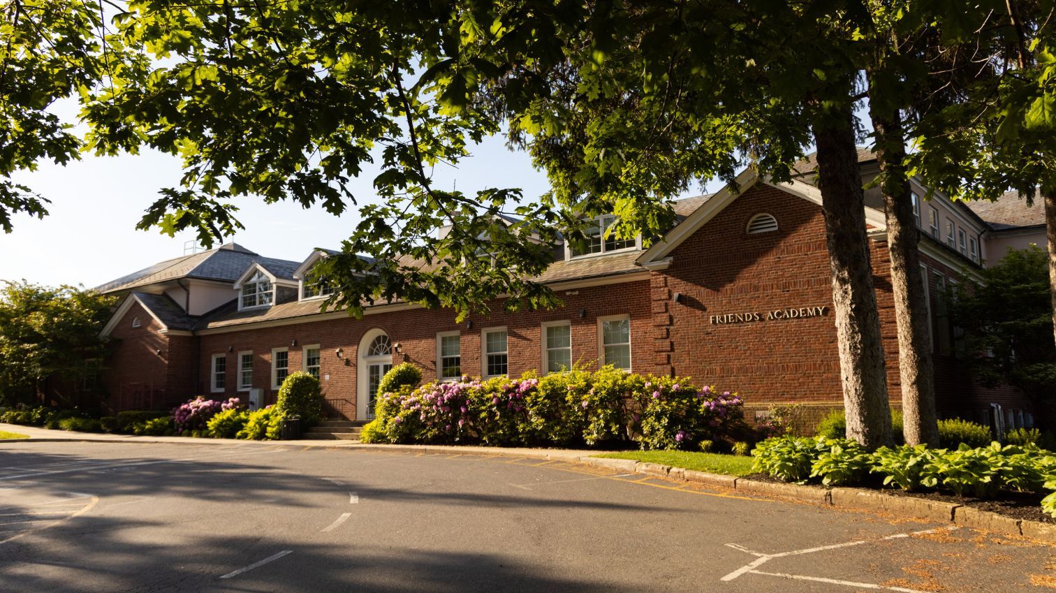 A brick school building with white-trimmed windows, flowering bushes, and trees in front. The sign on the building reads 