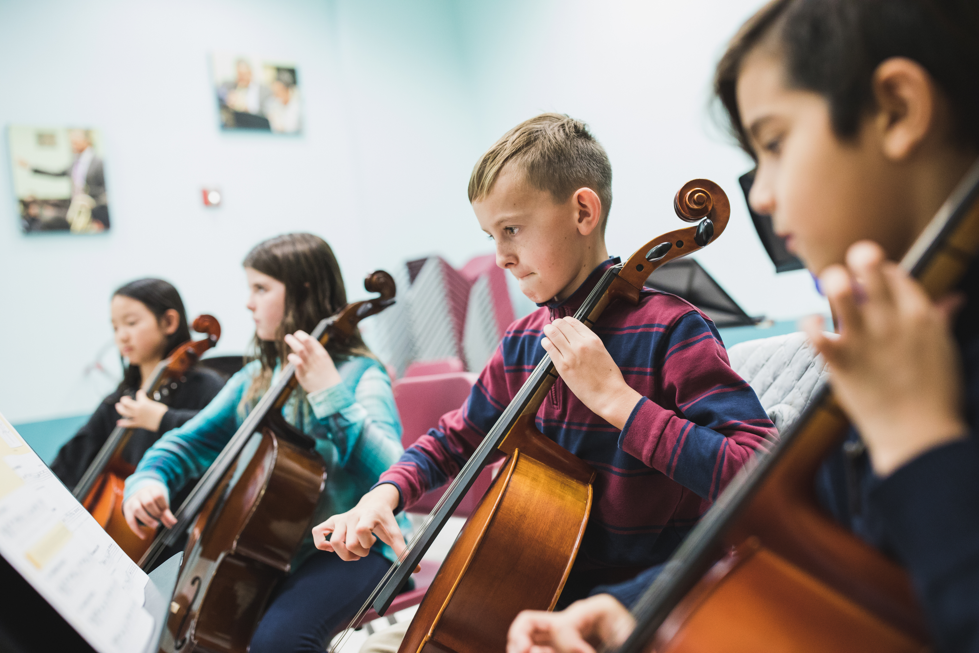 Four children sit in a row playing cellos in a classroom, focusing on sheet music in front of them.